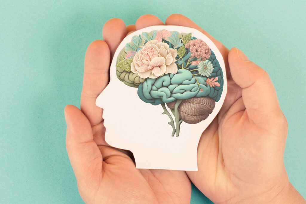 two hands holding a paper cutout in the shape of a human head, with the brain illustrated in detail as a collection of leaves and flowers